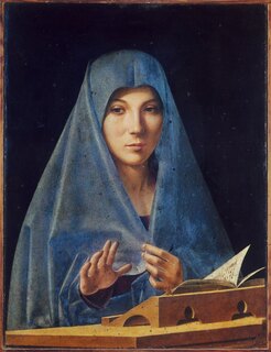 Woman in blue veil, one hand raised in benediction, the other holding the veil closed. She sits at a desk with a small wooden stand holding an open book.